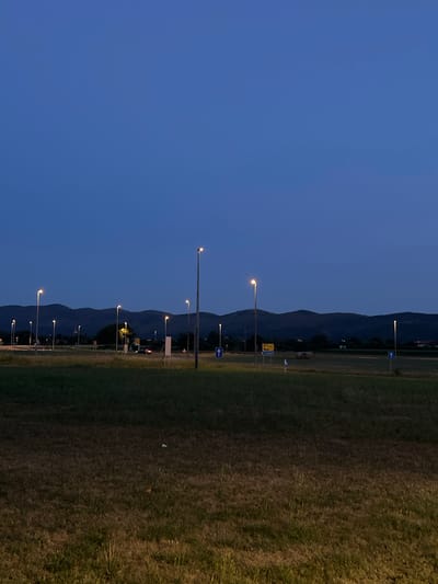 The image depicts a scene at dusk in Šempeter pri Gorici, Slovenia, likely an open area near a road or intersection. A vast expanse of dry grass dominates the foreground, with some areas appearing darker and shadowed, suggesting uneven terrain or a transition from daylight to night. Scattered across this grassy field are numerous tall, slender lampposts, many of which are illuminated, casting a warm glow on their surroundings.

In the mid-ground, a road or pathway is visible, lined with more lampposts. A single car appears on this road, its headlights on. Several road signs are present, including yellow directional signs with text and blue arrow signs. In the distance, a line of dark, rolling hills or mountains forms the horizon against a deep blue twilight sky. A few scattered trees are visible in the mid-ground, along with some large, cylindrical hay bales, suggesting the area might be adjacent to agricultural land. The overall atmosphere is calm and serene, characterized by the soft light of the lamps and the fading daylight.