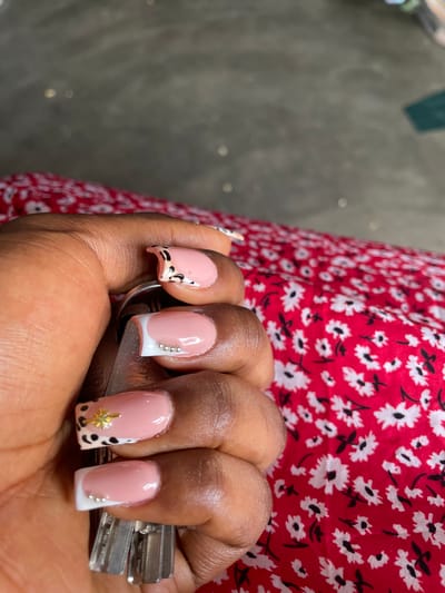 A close-up shot shows a person's hand holding a set of keys against a red fabric with a white floral pattern. The hand has several manicured nails with a pink base, white French tips, and some decorative elements including small silver beads and a gold star embellishment on one nail, and a leopard print design on another. The background is out of focus, showing a textured gray surface. The image was captured in Zanzi, Nigeria.