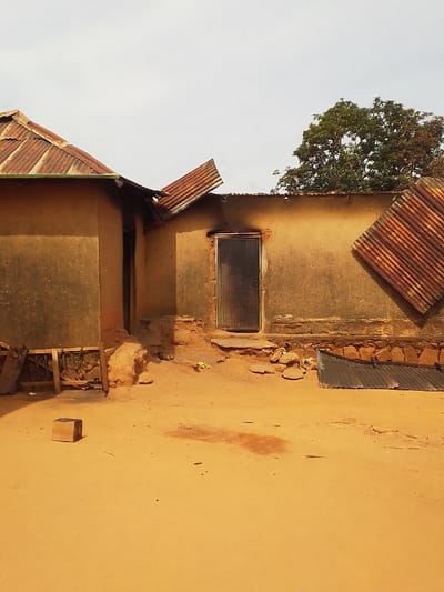 The image shows a partially destroyed building in Kwal, Nigeria, located at latitude 9.804 and longitude 8.646. The structure is made of mud brick and has a corrugated metal roof. Several sections of the roof are missing, and a large section of the roof is lying on the ground in front of the building. There is a single doorway leading into the building, but the door is closed. There is a small tree visible in the background to the right of the building. There is a sandy area in front of the building, littered with debris from the roof. There are no people present. The scene appears to have been damaged by a storm.