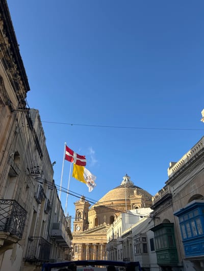 The image depicts a street scene in Mosta, Malta, under a clear blue sky, with the Rotunda of Mosta (Mosta Dome) as the central background element. The large, light-colored dome, topped with a smaller cupola, dominates the view. Its classical facade below features columns and a clock tower visible on the left.
Two flags fly from poles positioned left of the dome: a red flag with a white cross (Order of Malta) above a yellow and white flag with a central crest (Vatican City). Buildings line both sides of the narrow street. The building on the left is multi-story, constructed of light-colored stone, with black metal balconies and visible external utility wiring. Buildings on the right exhibit similar stone construction, including traditional enclosed wooden balconies, one painted green and another blue.
The upper portion of a blue open-top tour bus is visible at the bottom of the frame, containing the heads of several passengers. A single utility wire traverses the sky horizontally above the street. The scene is illuminated by direct sunlight.