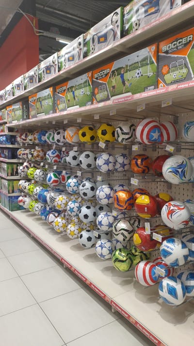The image depicts a retail display of soccer balls and soccer goal sets in what appears to be a store. The shelves are stacked with various soccer balls of different designs, colors, and sizes. Some of the soccer balls are branded with logos. Boxes of "Soccer Goal Set" are also visible on higher shelves, featuring illustrations of children playing soccer. The scene is well-lit, suggesting an indoor commercial environment. The retail setting is consistent with a toy store or sporting goods store. The floor is tiled with a light-colored material. Based on the signage, which includes Greek text "Ένας κόσμος όνειρο!" (meaning "A world of dreams!"), the location is likely a store in Greece. Specifically, based on the store's signage and product offerings, this image was taken at a Jumbo store in Pylaia Municipal Unit, Greece, which is located in the Thessaloniki regional unit.