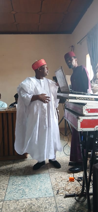 The image depicts two individuals in a room, possibly a place of worship or a cultural gathering in Sabon Gari North, Nigeria. The scene is illuminated by artificial light, suggesting it could be any time of day.

The foreground subject is a man dressed in a white traditional robe and a red and gold patterned cap. He is facing left, with his head tilted upwards, and his right hand is raised. A ring is visible on his right ring finger. His expression appears contemplative or devotional.

Behind him, slightly to the left, another man is visible, wearing a dark maroon robe and a dark red cap. He is also facing left, with his head turned downwards. His face shows a beard. He appears to be standing near or operating some equipment.

The background features musical equipment, including a large, red and silver case with the visible text "ODYUSA". There are also wires and a power outlet on the floor, indicating that the equipment is functional. To the far left, a wooden door or cabinet with decorative paneling is partially visible. The floor is tiled with a mosaic pattern of gray, white, and light blue. The overall impression is a candid shot during a performance or a religious ceremony.