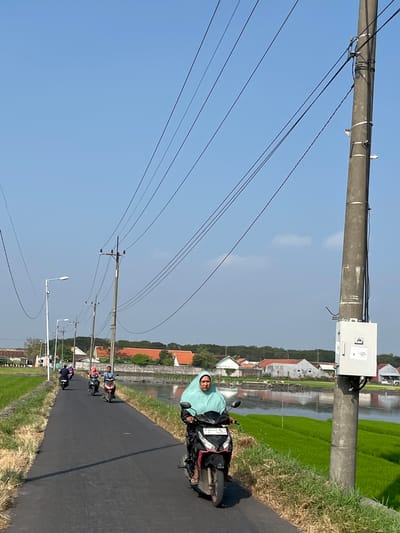 A medium shot captures a bright, clear day in Kalitengah, Indonesia, with a view of a rural road lined by rice fields. A woman in a turquoise hijab rides a black and red motorcycle in the foreground, facing the camera. Behind her, on the same asphalt road, four other motorcycles travel in the same direction, their riders largely obscured by distance and the vehicles themselves. The road is flanked on the left by a grassy verge and vibrant green rice paddies, and on the right by more rice fields and a body of water reflecting the sky. Utility poles with numerous wires stretch across the sky, casting shadows on the road. In the background, traditional Indonesian houses with red-tiled roofs are visible amongst trees and vegetation. A grey utility box is mounted on the closest utility pole. The sky is a clear, vibrant blue with a few wispy clouds.