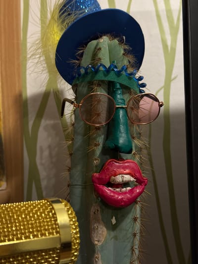 The image displays a whimsical and surreal indoor still life, centered around a pale green columnar cactus lying horizontally. The cactus is adorned with an array of humorous accessories, creating an anthropomorphic character.
On the left side of the cactus, a miniature dark blue sombrero is perched, featuring glitter on its brim and a fluffy yellow feather protruding from underneath. A bright blue spiral ornament is also attached near the hat. In the middle section of the cactus, a pair of round, rose-tinted sunglasses with delicate gold frames rests. Directly beneath the sunglasses and extending to their right are two small, dark green, textured objects, resembling stylized bottles or decorative elements. Further to the right, a striking bright pink object, shaped like exaggerated, open lips with rough, white tooth-like structures inside, is impaled onto the cactus spines. The cactus itself has prominent ribs and numerous sharp, light-colored spines.
In the bottom right corner of the frame, a golden microphone with a distinct mesh texture is partially visible, suggesting it's part of the surrounding environment or another artistic arrangement. The background features a light-colored surface, likely a wall, decorated with a subtle, abstract pattern of pale green, wave-like lines. A wooden surface is visible at the very bottom edge of the image.
No people are present, and no specific activity or event is taking place; the scene is a static arrangement. The lighting suggests an indoor setting, possibly under artificial light, without any indications of outdoor weather or specific time of day. The overall impression is one of playful creativity. There is no visible text in the image. The provided location context for this media file is Ново-Никольское, Russia.