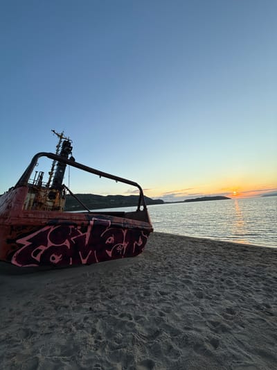 This image captures a sunset scene on a sandy beach in Teriberka, Russia. In the foreground, the rusted hull of a beached ship dominates the left side of the frame. The ship is covered in graffiti. The sand on the beach is textured with footprints. The ocean is visible in the mid-ground, with gentle waves reflecting the warm hues of the setting sun. The horizon is marked by a chain of hills and a faint outline of a town or structures. The sky transitions from a clear, pale blue at the top to a soft gradient of yellow and orange near the horizon, where the sun is setting. The overall atmosphere is serene and somewhat melancholic.