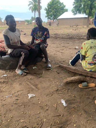 This image depicts a group of young people sitting on the ground in what appears to be an outdoor, possibly rural, setting in Dull A, Nigeria. The ground is a mixture of dirt and sand, with scattered leaves, twigs, and some debris, including small pieces of plastic. In the background, there is a building with a corrugated metal roof and a brick wall. Lush green trees are visible on the left side of the image, suggesting a somewhat natural environment.

There are at least four individuals visible, though their full bodies are not always in frame. Two of the individuals are more prominently featured in the foreground, appearing to be conversing or interacting. One person is wearing a light-colored t-shirt with a graphic design and the text "JUNIOR RGACMEN" partially visible. Another individual is wearing a dark blue t-shirt with a colorful graphic. A third person is seated behind them, wearing a yellow t-shirt with a graphic and text that includes "BO" and possibly other obscured letters. The fourth person's clothing is mostly out of frame, but they are wearing dark pants and a t-shirt.

The individuals are dressed casually, and their postures suggest a relaxed atmosphere. One person is wearing blue and white striped pants and light blue sandals, while another is wearing dark pants with white stripes and light blue sandals. A third person has a brown sandal to their right, and another dark sandal is visible near the foreground.

The lighting suggests it is daytime, possibly mid-morning or afternoon, with diffused light and no strong shadows, indicating overcast weather or a shaded area. The overall scene conveys a sense of everyday life and social interaction among young people in a communal outdoor space.