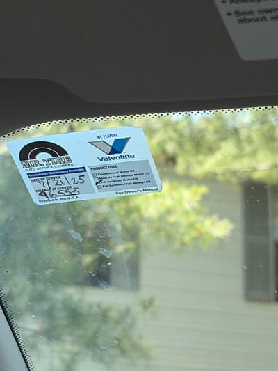 An interior shot from a car shows a maintenance sticker affixed to the windshield, set against a blurred outdoor background. The sticker, primarily white and blue, is from "MR. TIRE AUTO SERVICE CENTERS" and features the "Valvoline." logo, indicating a "Preventative Maintenance Reminder." Under "PRODUCT USED," "MaxLife High Mileage Motor Oil" appears to be checked. The "DATE OF SERVICE" is clearly marked as "7/21/25", and the "MILEAGE AT SERVICE" is "96555". The sticker also states "Printed in the U.S.A." The windshield exhibits various smudges and residue. To the left, a dark gray, textured car interior component, likely a sun visor or headliner, is visible. Partially obscured text on another label can be seen on this component, reading parts of phrases such as "• See ow" and "about a". Through the windshield, a daytime scene is visible with blurred green foliage and a light-colored building in the background. The image is taken in Lawrence Township, United States of America.