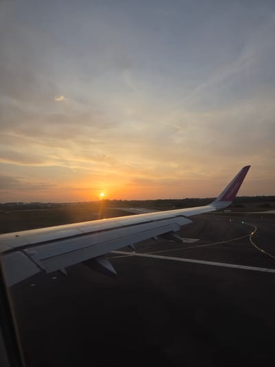 The image shows a view from an airplane window during sunset. Part of the plane's wing is visible, with the website "wizzair.com" printed on the winglet. Below the wing, the ground is visible, suggesting the plane is either taking off or landing. A runway with lights and markings is visible on the ground. The sun is setting on the horizon, casting a golden glow across the sky and land. The location is North Hertfordshire, United Kingdom, as identified by the airport environment and airline details. There are no people directly visible in the image. The focus is on the sunset and the airplane wing.