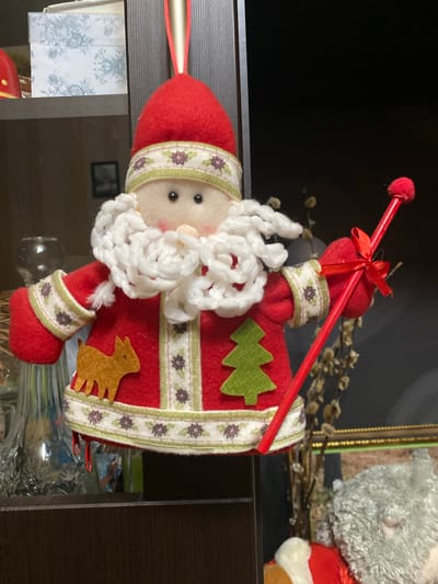 This is a medium shot, eye-level, indoor photograph of a handmade Christmas ornament of Santa Claus. The ornament is made of red felt and features white yarn for Santa's beard and mustache, black bead eyes, and a pink felt nose and cheeks. Santa is wearing a red hat with a white band adorned with a green border and small purple flowers. His red coat has a similar white band with purple flowers and green trim. A small, golden felt reindeer is affixed to the left side of his coat, and a green felt Christmas tree is on the right side. Santa is holding a red stick with a red pom-pom on top, adorned with a red ribbon bow. The ornament is hanging from a red ribbon. The background is dark and out of focus, with hints of shelves and other decorations, including a large glass vase on the left and what appears to be dried branches and another stuffed figure on the right. There are no people visible in the frame. The scene is consistent with a home or store interior, likely decorated for the Christmas holiday. There is no information within the image to suggest a specific location in Yokohama, Japan.