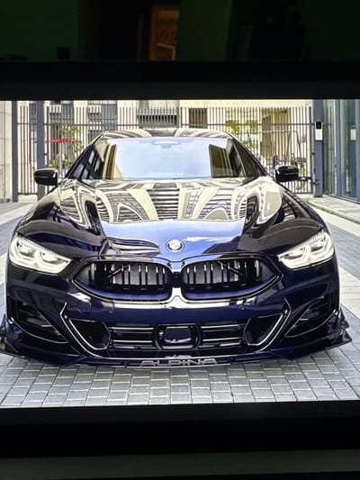 This is a low-angle, head-on, full-shot, indoor photograph of a dark blue, high-performance sedan parked on a light gray, brick-paved driveway. The vehicle, which appears to be a BMW Alpina, is the primary subject. It is clean and polished, reflecting the surrounding environment. The car's front grille, headlights, and aggressive front bumper are clearly visible. The background consists of a metal fence and a building with large windows, suggesting an urban or commercial setting. The image is captured from a slightly elevated perspective, looking down at the car. There is no discernible information about the location being Sant'Angelo di Piove di Sacco, Italy, nor are there any people present in the image.