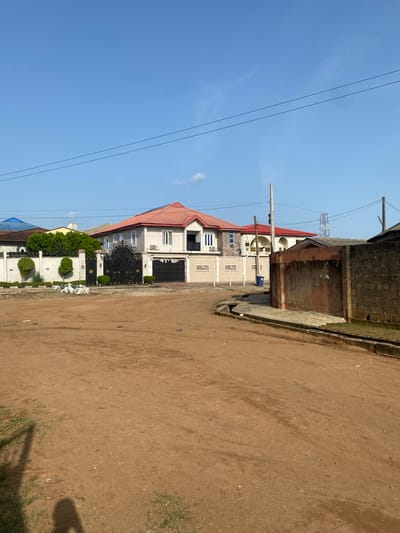 The image shows a street scene in Idimu, Nigeria. The road is unpaved, consisting of brown dirt. The right side of the image shows a partial view of a building constructed of brown bricks. In the background, there are two houses visible. One is cream-colored with a red roof, and a black gate. The other building is partially visible, with a blue roof. Power lines run across the top left of the image against a clear blue sky.