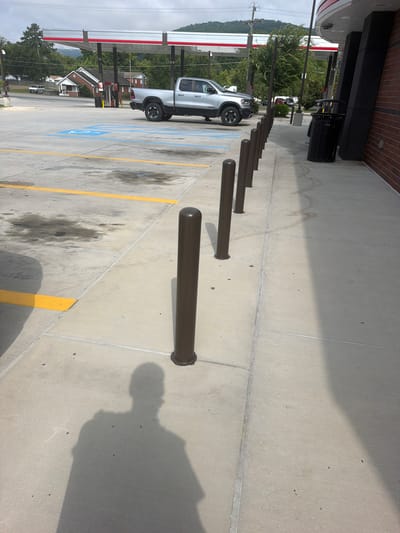 The image shows a sidewalk outside a gas station in Chattanooga, Tennessee. In the background, a silver pickup truck is parked at a gas pump under a red and white canopy. The sidewalk is lined with dark brown bollards. A black trash can is visible near the entrance of the building. The ground is concrete and shows some oil stains. The shadow of a person is cast on the sidewalk. The sky is cloudy, and trees and buildings are visible in the distance.