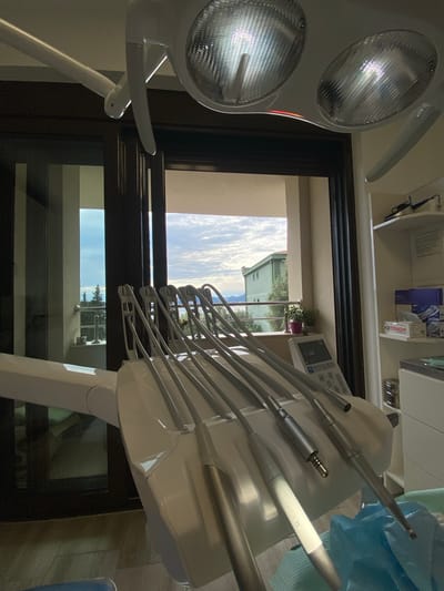 The image depicts the interior of a dental office in Podličak, Montenegro, as evidenced by the outdoor view visible through the window. The primary focus is on dental equipment, including a dental examination light and a dental unit with various instruments attached. The dental unit holds several silver-colored instruments, including a white-handled instrument. Outside the window, a building with a red-tiled roof is visible, along with a sky filled with clouds. The inside shelves can be seen with medical supplies. The equipment's modern design suggests a contemporary dental practice. The composition of the photo indicates it was taken from the perspective of someone situated within the office, likely a patient or dental professional.