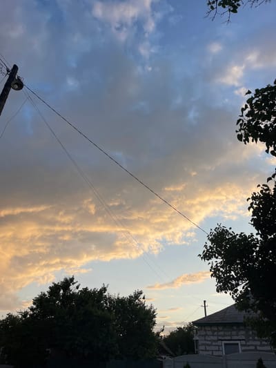 A low-angle, medium shot captures a dusky sky filled with clouds tinged with orange and yellow from the setting sun, contrasting with patches of blue. A wooden utility pole with a light fixture stands on the left, supporting taut electrical wires that stretch diagonally across the frame. Silhouetted trees frame the top right and the foreground. In the lower right quadrant, the roof of a white brick house, partially constructed and featuring a dark-trimmed window, is visible behind a white fence. The scene appears to be set in a suburban or rural area of Krasnokutsk, Ukraine.