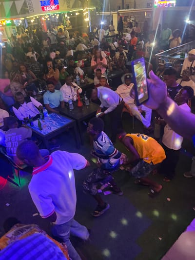 The image depicts a vibrant outdoor gathering at "JULETION RESORT" in Oba, Nigeria. The scene is illuminated by colorful lights, creating a lively atmosphere. A crowd of people, dressed in casual and festive attire, are seen mingling, some seated at tables with drinks, while others are standing and engaged in conversations. In the foreground, several individuals are actively participating in a dance or celebratory activity, with some in motion and others observing. There are tables with bottles of drinks and what appear to be water bottles scattered on the ground, suggesting a lively event. The resort's signage is prominently displayed, identifying the location. The overall impression is one of a social event or party in full swing.