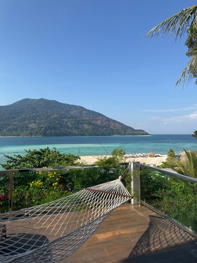 The image presents an elevated, panoramic view of a tropical coastal environment in Ban Ko Li Pe, Thailand, under a clear blue sky. In the immediate foreground, a white, rope-net hammock with wooden spreader bars is suspended on a dark wood-paneled balcony. The balcony is bordered by a railing constructed of stainless steel posts and glass panels. Sunlight, originating from the right, casts distinct shadows of the hammock netting and railing onto the balcony floor. Lush green and yellow flowering foliage is visible just beyond the railing.
The midground features a curved white sand beach, bordered by a calm sea exhibiting distinct bands of turquoise in the shallows and deeper blue further offshore. Numerous white beach umbrellas and sun loungers are arranged on the sand, alongside a few small, thatched-roof structures. Several small boats are visible in the water. A large, heavily forested island or mountain dominates the background, stretching across the horizon. A portion of a palm frond is visible in the upper right quadrant of the image. No individuals are present within the frame.