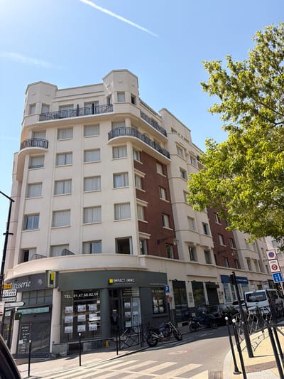 This is an eye-level, outdoor, daylight shot of a building facade on a street in Courbevoie, France. The building is modern, with a cream-colored facade interspersed with sections of red brick. It features numerous windows, some with balconies. A large tree with green leaves overhangs the top of the building from the left side of the frame. On the street level, there is a storefront for "IMPACT-IMMO," a real estate agency, with "L'IMMOBILIER, AUJOURD'HUI" written beneath it, along with a website address. Another storefront appears to be a bakery, labeled "Patisserie Au Paradis du Pain." A street sign indicates directions to "SEINE," "PUTEAUX," and "ARDIERES." Vehicles, including a white van labeled "AMBULANCES ZEPHYR," a black car, and two motorcycles, are parked on the street. Traffic signs, including a "ZONE 30" sign, are visible. The sky is clear and blue with a faint contrail. The overall scene suggests a typical urban environment on a sunny day, likely in the morning or afternoon given the lighting.