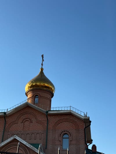An upward, angled shot shows the golden dome and cross of a Russian Orthodox church in Borzya, Russia, juxtaposed against a clear blue sky. The church's brick facade features decorative brickwork and arched windows. Metal railings and green roofing elements are visible. There are no people visible in the image.