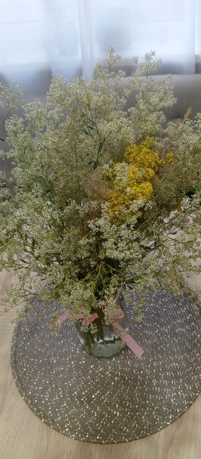 A bouquet of delicate white and yellow wildflowers sits in a clear glass vase on a round, textured placemat. A pale pink ribbon is tied around the neck of the vase. The placemat rests on a wooden surface. In the background, sheer white curtains hang against a neutral wall. The scene is brightly lit, suggesting it is indoors during the daytime. The location is a private residence in Petukhovo, Russia.