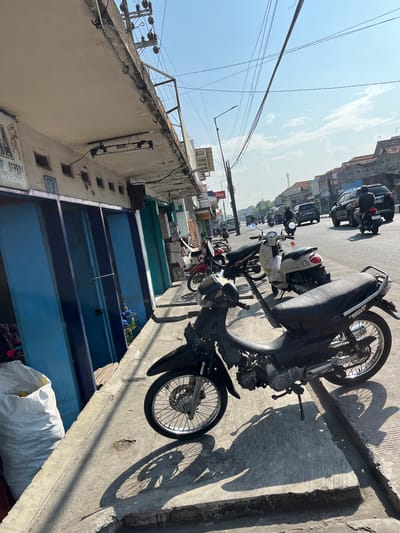 The image depicts a street scene in Gedang, Indonesia, on a bright, sunny day. In the foreground, a black motorcycle is parked on a concrete sidewalk. Its shadow stretches across the pavement. Behind the motorcycle, several other motorcycles and a white scooter are visible, also parked on the sidewalk.

The sidewalk runs alongside a row of shops with blue and green facades. A large white sack is placed on the sidewalk near the shops. Above the shops, a concrete overhang is supported by pillars, and electrical wires crisscross overhead.

The street itself is paved and features traffic, including cars and motorcycles with riders wearing helmets. Buildings with tiled roofs line the opposite side of the street, suggesting a commercial or residential area. The sky is clear blue with a few wispy clouds. The overall impression is that of a typical bustling street in an Indonesian city.