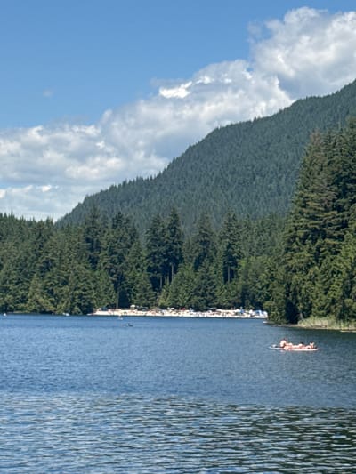 This is a vertical, eye-level, medium shot of a serene lake surrounded by dense evergreen forests and a majestic mountain. The water in the foreground is a deep blue with ripples, reflecting the clear sky and white, scattered clouds above. In the middle ground, the lake stretches towards a sandy beach crowded with people sunbathing and relaxing. Further back, the shoreline is lined with a dense wall of tall, dark green evergreen trees, with the lower slopes of a large, tree-covered mountain rising dramatically behind them. On the lake, a small raft is visible in the middle right of the frame, carrying several people lounging on it. The scene is bright and appears to be a sunny day. This view is characteristic of Buntzen Lake in Anmore, BC, a popular recreational area for Port Moody residents.