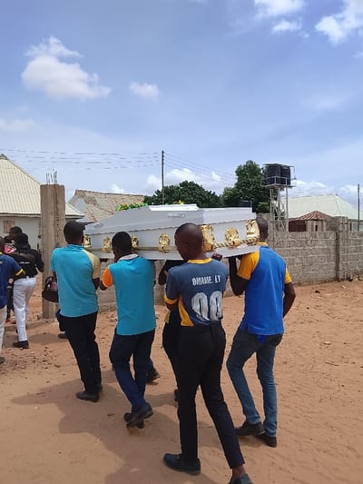 Six men are carrying a white casket with gold trim, while walking on a dirt road. They are walking in a line, and the casket is being carried by the middle four men. The men are wearing various casual clothes, including a blue shirt with a yellow trim, a blue and yellow shirt with the number 00, a turquoise blue shirt, a blue shirt, and a yellow shirt with the name "OMAME. LT". The men are walking away from the camera, towards a building in the background. The environment is dusty and dry, with a clear sky and a few clouds. The scene is likely in the town of Mukaiya, Nigeria, as the coordinates provided (8.4 latitude, 8.342 longitude) are located in the region. This scene likely depicts a funeral procession.