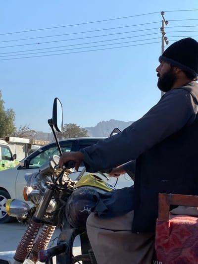 An outdoor, daytime scene in کوکران, Afghanistan, depicts an adult male seated on a motorcycle, facing right in profile. The man wears a dark knitted beanie, a dark long-sleeved shirt, and a dark vest over light-colored trousers, with a dark beard. His hands are positioned on the chrome handlebars of the motorcycle, which features a yellow fuel tank and a dark front section with the Arabic script "مهتر" (Mehtar) visible. A chrome side mirror extends from the handlebars. In the background, under a clear blue sky, multiple overhead power lines are visible, supported by a utility pole. Distant mountains are present on the horizon. Partially visible behind the motorcycle are other vehicles, including a white van and a light-colored sedan. Sparse trees and indistinct structures are also present in the background. A red, fabric-covered object is partially visible on the right side of the frame, behind the man.