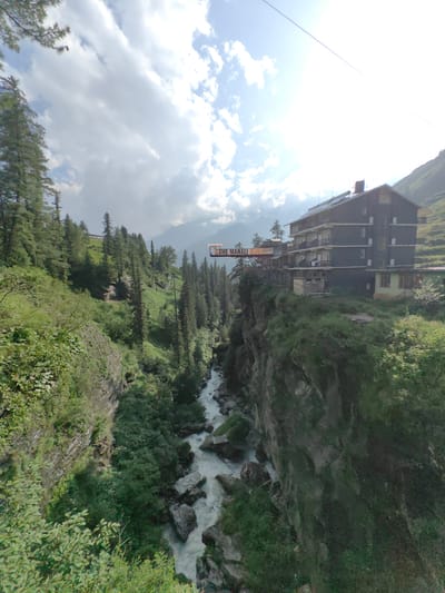 A building labeled "THE MANALI" sits precariously on the edge of a cliff overlooking a rushing river in Kothi, India. The multi-story building, painted dark blue, has several balconies and windows. A red bridge connects to the building, spanning a section of the cliff face. The surrounding environment is lush with green trees and vegetation, typical of a mountainous region. The sky is partly cloudy, with the sun shining through, creating bright patches. The river below flows rapidly over rocks, carving its way through the gorge. The overall scene depicts a dramatic landscape where human construction is integrated into a rugged natural setting.