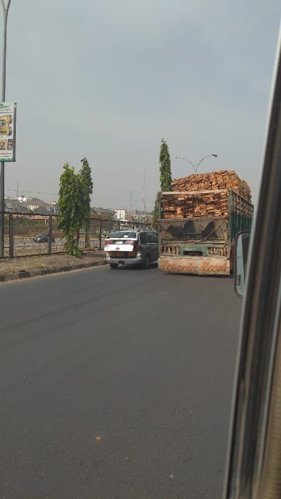 The scene depicts a multi-lane asphalt road in the city of Aso, Nigeria, viewed from inside a moving vehicle. The sky is a uniform, hazy light grey.

In the midground, a dark green truck, possibly a flatbed or stake-side model, is heavily laden with stacked light-colored wooden planks or processed timber, piled significantly above its cab height. The rear of the truck displays faded paint and partially visible text on a lighter-colored section, possibly "Nigeria Ruah". To the immediate left of the truck, a silver or light grey minivan travels with its rear hatch open, revealing the cargo area and a white placard or license plate.

To the left of the road, an unpaved median strip features sparse dry vegetation and two tall, slender trees with green foliage. Beyond this, a dark metal fence separates the primary road from a lower-level road where a dark-colored sedan or SUV is visible. Further to the left, a tall metal utility pole supports a green and white billboard displaying commercial advertisements, including what appears to be a "Shell" logo and product images related to lubricants or fuel. In the distant background, urban structures including buildings and communication towers are partially obscured by atmospheric haze. No individuals are explicitly visible in the image.