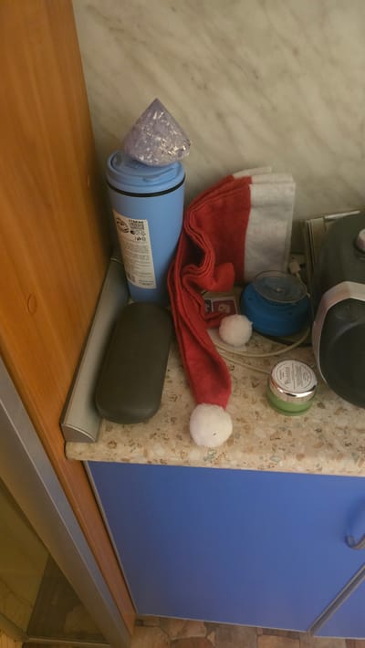 The image depicts a cluttered shelf or countertop. On the left, a wooden cabinet is visible. A light blue cylindrical container with some text on its side sits on the surface. On top of the blue container is a faceted, translucent purple object. To the right of the blue container, a red Christmas hat with white pom-poms is draped, and to its right, a stack of white folded items, possibly towels or cloths. A small blue circular object with a grid pattern is next to the hat, and behind it, wires are visible. Further to the right, a green circular container with a silver lid is present, and a dark grey object, possibly a speaker, is on the far right. The surface appears to be made of a speckled material. A blue cabinet is visible below the shelf. The scene does not provide any information that would allow for the determination of its location in Ukhta, Russia.