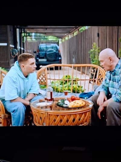 Two men are seated at a round, wicker table outdoors. The man on the left is younger, with short, light brown hair and is wearing a light blue hooded sweatshirt and blue jeans. He is looking to the right. The man on the right is older, bald, and is wearing a blue and green plaid shirt and brown pants. He is looking towards the table and appears to be speaking. On the table, there is a bottle of dark liquid, two small glasses, three red tomatoes, and a piece of flatbread or pastry. Behind them, a wooden fence and lush green foliage are visible. A dark-colored SUV is parked in the background, under a covered area. The scene appears to be a residential backyard or outdoor seating area, possibly in Ivanovo, Russia, given the context.