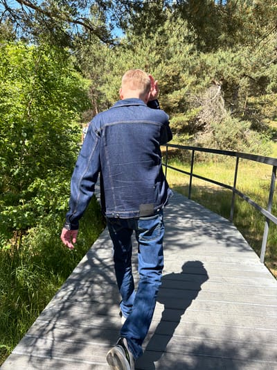 The image shows a person walking on a wooden boardwalk through a wooded area with green foliage and tall pine trees. The person is wearing a dark blue denim jacket and jeans. The boardwalk has a metal railing on the right side. Sunlight filters through the trees, casting shadows on the boardwalk. The location appears to be a nature trail or park. Based on the provided context, the location is identified as the city of Morskoye, Russia.