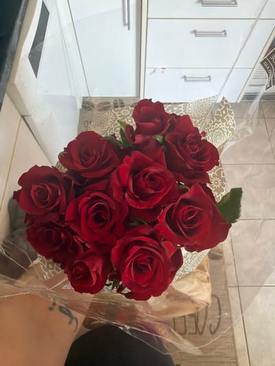 A bouquet of deep red roses is the focal point of the image. The roses are arranged in a dense cluster, and they are partially wrapped in patterned paper and clear cellophane. The bouquet rests on a white, smooth surface. In the background, there is a white cabinet with silver handles. The background is slightly blurred, and the wall appears to be beige. Part of a person is visible on the right side of the picture. A tattoo is visible on the person's skin. The location appears to be indoors, likely a kitchen or similar space. There is nothing in the image that indicates the location is in Rapperswil-Jona, Switzerland.