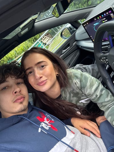 A couple poses in a car for a selfie. The car's navigation screen shows an address in Llangynog, United Kingdom. The woman has long, dark brown hair and is wearing a camouflage jacket. The man has curly, dark brown hair and is wearing a blue and gray hoodie with a red and white logo. They are both looking at the camera and smiling slightly. The car's interior is modern and sleek. Outside the window, greenery is visible.