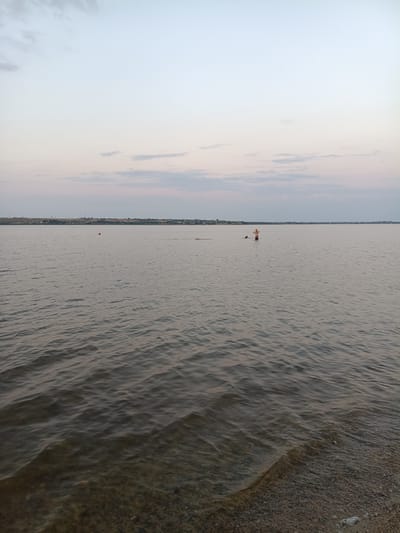 A wide-angle, eye-level shot captures a serene body of water, likely a lake or river, on the outskirts of Nezavertaylovka, Moldova, during twilight. The sky is a gradient of pale blue to light gray, with faint streaks of pink and scattered clouds, suggesting the sun has recently set or is about to rise. The water's surface is calm, with gentle ripples reflecting the muted sky colors.
In the distance, a long, low-lying horizon line features a sparsely populated shoreline with scattered trees and buildings, indicative of the village. A red buoy floats in the middle distance, its bright color a small contrast to the subdued tones of the landscape. Closer to the center of the frame, two individuals are visible in the water. One person, appearing to be standing, has their arms raised. Another person is partially submerged. To the left of these individuals, another small red marker is visible in the water. The foreground shows the edge of a shallow, pebbly shore where the water gently laps.