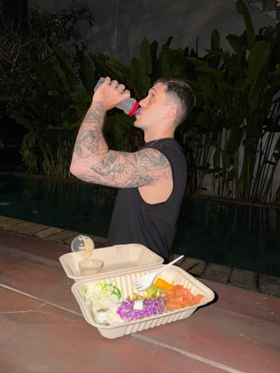 A man with extensive arm tattoos drinks from a clear water bottle with a red cap. He is wearing a black sleeveless shirt. In front of him, on a wooden table, are two takeout containers. The closer container holds a salad with salmon, orange pieces, purple cabbage, and green lettuce. The second container, slightly behind and to the left, holds a dip. The background shows a dark swimming pool and lush green foliage. The scene appears to be outdoors at night, possibly in Kerambitan, Indonesia, given the tropical vegetation.