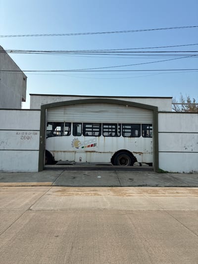 A parked, white bus with the logo "Coatzacoalcos 2000" is partially visible inside a garage. The garage door is arched and the garage is marked "M. ANGEL DE QUEVEDO 2608". Overhead power lines are in the upper left of the frame. Concrete sidewalk and road are visible on the right side of the garage.