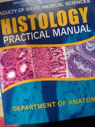 The image displays the cover of an educational textbook titled "HISTOLOGY PRACTICAL MANUAL." The book cover features a dark blue background with prominent yellow lettering and accents. The main title "HISTOLOGY" is presented in large, bold yellow capital letters, with "PRACTICAL MANUAL" below it in smaller, grey uppercase text.

Above the main title, partially visible text identifies the institution: "NNAMDI AZIKIWE" (partially obscured) "COLLEGE OF HEALTH SCIENCES" (partially obscured), and "FACULTY OF BASIC MEDICAL SCIENCES." At the bottom of the cover, also in yellow uppercase letters, "DEPARTMENT OF ANATOMY" is visible (partially obscured).

A prominent yellow wavy line horizontally divides the cover, separating the title area from a central band featuring three distinct microscopic images. These images, stained predominantly in pink and purple, depict various biological tissues and cellular structures, consistent with the subject of histology. From left to right, the images show what appear to be kidney tubules, glandular tissue, and potentially seminiferous tubules or similar structures.

The setting is an academic or medical environment, with the book serving as a practical guide for scientific study. The context provided indicates the location is Otu, Nigeria. No individuals are present, and there are no discernible details regarding the time of day, weather, or emotional context. The focus is entirely on the book's design and its educational content.