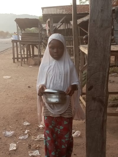 A young girl in a white hijab stands in a dusty, impoverished setting. She wears a striped shirt and a long skirt with a red and blue pattern. She holds a small, shiny metal container, possibly for food. Litter is scattered on the ground around her. Behind her are makeshift wooden structures, including market stalls. In the background, a road and distant hills can be seen under a hazy sky. The overall impression is one of poverty and hardship.