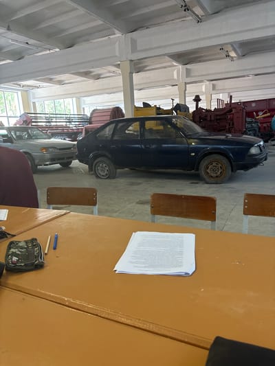 In an indoor industrial or workshop setting, a dark blue sedan is parked in the foreground. The vehicle appears aged, with visible rust on the wheels and a flat rear tire. Behind the sedan, a light-colored sedan is partially visible, facing the same direction. Further in the background, various agricultural or industrial machinery, some painted red and yellow, are present. The space is characterized by a concrete floor and a ceiling with exposed beams and lighting fixtures. In the immediate foreground, a brown wooden table is visible, with a small camouflage pouch, a pencil, and a blue pen on its surface. A stack of documents is also placed on the table. The overall scene suggests an indoor storage or maintenance area for vehicles and machinery.