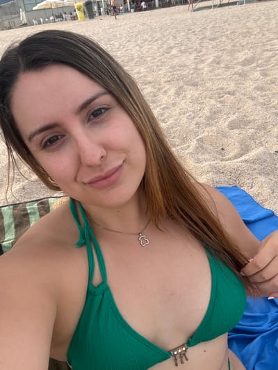 A young woman with long, light brown hair is pictured on a beach in Vilassar de Mar, Spain, with the Mediterranean Sea visible in the background. She is lying on a blue towel, wearing a green bikini top and a delicate necklace. The sand on the beach is light in color and scattered with small shells. In the background, several beach umbrellas, tables, and chairs suggest a beachfront restaurant or bar. People are visible in the distance, some walking along the shore and others seated at tables. The woman is looking at the camera, with her head tilted slightly and a gentle smile on her face.