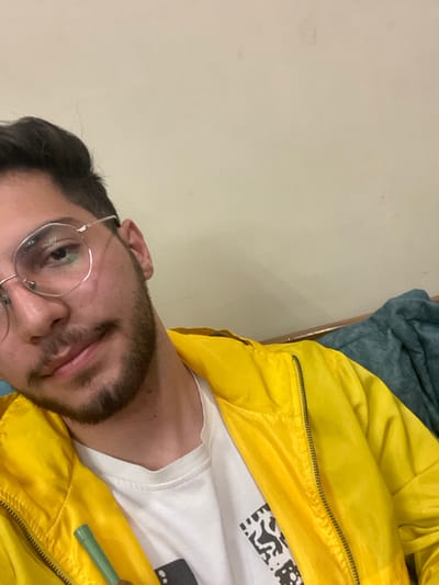 The image displays a head and upper torso shot of a young adult male in an indoor setting. The male subject has dark, short hair, a trimmed beard, and a mustache. He wears round, thin metal-rimmed glasses and looks directly at the camera with a slight smile. He is dressed in a bright yellow zippered jacket over a white t-shirt that features a partial black and white graphic design. A small, light green cylindrical object, possibly a plastic straw or similar item, is visible in the lower left portion of the frame, held near his chest. The background consists of a plain, light-colored wall, with a horizontal wooden edge and a folded blue-green fabric visible in the upper right. The specific location within Homs, Syria, cannot be determined from the visual information presented.