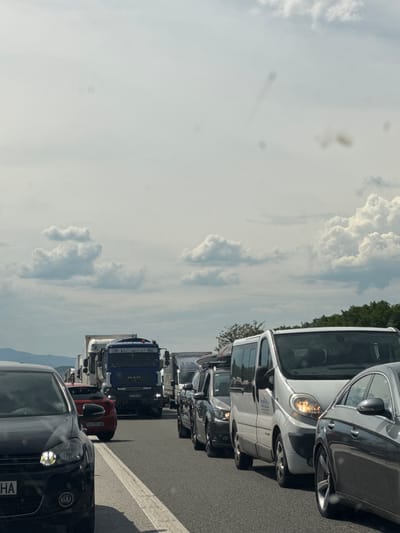 This media file shows a line of vehicles stopped on a highway, indicating a traffic jam. The scene is viewed from the perspective of a car passenger looking forward.
The subjects include various vehicles such as cars, vans, and a large truck. The truck is blue and has "MAN" written on its front. Other vehicles include a silver van with visible text on its side, a black car in the foreground with its headlights on, and a red car behind it. The setting is a highway with a white line marking the lanes and a shoulder with tire tracks. In the background, there are trees and a cloudy sky.
The activity taking place is traffic congestion, with vehicles stopped or moving very slowly. The time of day appears to be daytime, likely afternoon, given the brightness and the shadows. The weather seems to be partly cloudy, with large white clouds visible in the sky. There are no people prominently visible in the scene, except for possibly blurred figures inside the vehicles.
Notable details include the license plates on some of the vehicles. The red car has a license plate "EA 1991 AB". The black car in the foreground has a partial license plate visible: "HA". The silver van has text on its side that includes "AUTO-POL" and "PREFABRICATED". The overall mood conveyed is one of typical traffic, possibly with a hint of frustration due to the delay. The location context provided is Shtarkovo, Bulgaria, which suggests the highway is in or near that region.