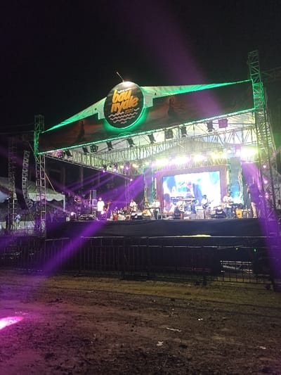 The image depicts an outdoor music festival or concert taking place at night. A large, brightly illuminated stage, constructed with metal scaffolding, is the central focus. A prominent banner stretches across the top of the stage, featuring a circular, glowing logo that clearly displays "Festival Bau Nyale 2023." The banner also shows stylized figures, possibly dancers, on either side of the central logo.
On the stage, a band is performing. Multiple musicians are visible, playing instruments such as guitars, drums, and keyboards. Several band members, dressed mostly in white shirts, are actively engaged in the performance. A large LED screen at the rear of the stage displays an image or video, likely related to the performance. The stage is extensively lit with numerous spotlights and LED fixtures, emitting bright white, green, and purple light beams that cut through the dark night. Notably, distinct purple light rays illuminate the foreground.
In front of the stage, metal barricades separate the performance area from the audience space. The ground in the immediate foreground appears uneven and muddy or dusty. In the far background, to the left of the stage, a white tent or structure is partially visible. The scene conveys an energetic and lively atmosphere, characteristic of a live music event. The event is identifiable as the "Festival Bau Nyale 2023," taking place in Kecamatan Pujut, Indonesia.