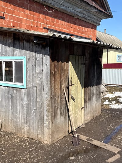 The image presents an external view of an old, weathered wooden structure, possibly a shed or outbuilding, located in what is presumed to be Кипчак, Russia. The structure features a small window with a light blue frame. To the left of the wooden structure, there is a red brick chimney or wall. The roof is corrugated and made of gray metal. The wall is constructed of old wooden planks of different colors, which were exposed to the sun and weather. A shovel is leaning against the exterior, a section of which has been painted yellow. The ground surrounding the structure is dirt with bits of debris scattered around. There is snow and ice in the background along with a building in the distance that is painted yellow and red. The sky is clear and light blue.