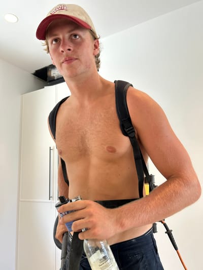 A fair-skinned person with a cap poses indoors. The person wears a black harness over their bare chest. They are holding a glass bottle and a blue tube-like object. White cabinets are visible in the background. The person has short, curly hair and is looking directly at the camera. The image is taken from a slightly low angle and rotated.
