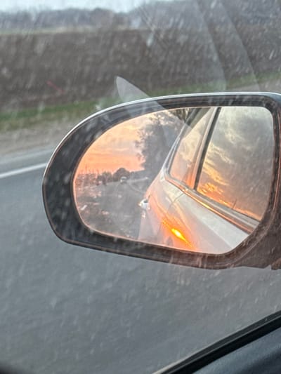 The image displays a view from inside a vehicle, focusing on the driver's side exterior mirror, observed in Krasnoe Pole, Russia. The side mirror, framed in black, shows surface dirt and reflections. Within the mirror's reflection, an orange and pink sunset or sunrise sky is visible above a distant tree line and a white vehicle, appearing to move away from the observer. An illuminated amber turn signal on the side of the vehicle from which the photograph is taken is also distinctly reflected in the mirror. Outside the mirror, through the vehicle's window, a gray road surface is visible to the left, and a blurred, dark line of trees or vegetation extends in the background under an overcast sky. No individuals are depicted. The scene suggests vehicular travel during dawn or dusk.