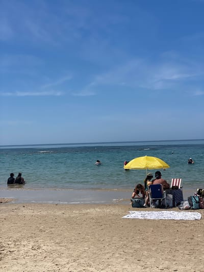 The image depicts a scene at a beach in the city of  Mihmoret, Israel, located at latitude 32.408 and longitude 34.868. Four individuals are gathered beneath a yellow umbrella, situated on a sandy beach. Three of them are seated while one, a woman in a black bikini, is kneeling on the sand in front of a blue cooler. Two of the seated individuals, one male and one female, are facing the ocean, while the other seated individual is facing the opposite direction, obscured by the umbrella. The scene is characterized by a clear blue sky, calm turquoise waters, and a sandy shoreline. There are several people in the water, and the scene is tranquil and peaceful.