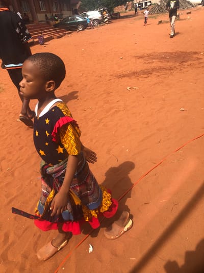 A child lies on their side on the red dirt ground in Nteje, Nigeria. The child is wearing a colorful, patterned dress and tan-colored sandals. An adult arm reaches into the frame from the left, and other people and buildings are visible in the blurred background. An orange cord lays on the ground near the child.