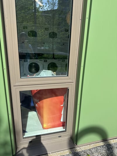 A ground-level, eye-level shot captures a windowed exterior wall, likely of a commercial or service establishment. The wall is painted a light green color with a horizontal dark beige trim supporting two windows. The left window reflects an interior view of a laundromat with multiple washing machines labeled with alphanumeric codes such as "D4," "D5," "W2," "W3," and "W4." The reflections on the window surface are superimposed on the interior view, with tree foliage and light reflecting off the washing machines. The right window reflects an indoor scene with a partially visible red and white promotional advertisement sign. The bottom right corner of the frame reveals a section of light-colored concrete or paving stones. The location is speculated to be in Hull, United Kingdom.